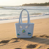 Light blue rubber tote with angel wing, whale and globe charms standing on sand by the shoreline