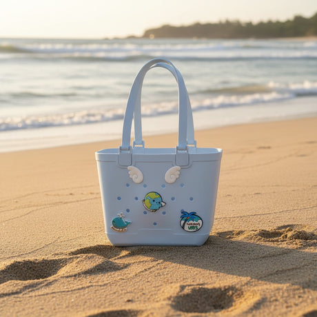 Light blue rubber tote with angel wing, whale and globe charms standing on sand by the shoreline