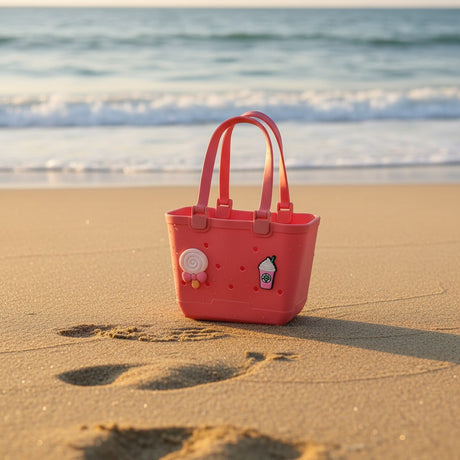 Coral pink waterproof beach tote with lollipop and drink charms standing on wet sand near waves
