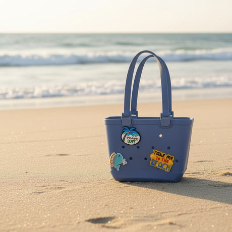 Navy blue sand proof tote with palm tree, whale and slogan charms standing on damp sand near the waves