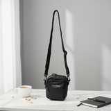 Small black crossbody bag on a white surface with a cup and notebook in the background