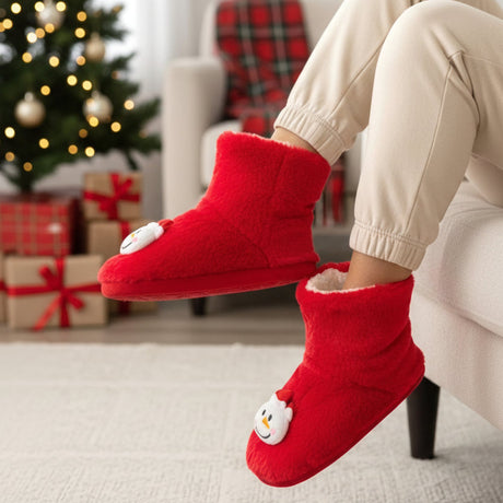 Red plush bootie slippers with a white snowman patch worn beside a Christmas tree