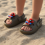 Kids olive green slides with red Spiderman charms, worn barefoot on soft beach sand