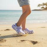 Lilac foam clogs with ventilation holes kicking up sand mid stride