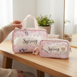 Pink 'Hello Spring' travel bag and pouch on a wooden table with a plant in the background