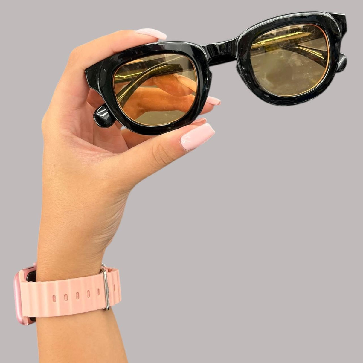 Hand holding a pair of black stargaze sunglasses with gold reflective lenses against a grey background