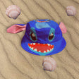 Blue Stitch character bucket hat with pink ears lying flat on sand between seashells
