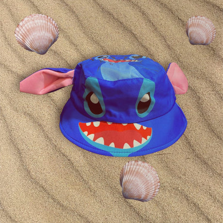 Blue Stitch character bucket hat with pink ears lying flat on sand between seashells