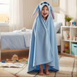 A young child stands barefoot in a bright bedroom, wrapped in a light blue hooded blanket with long bunny ears and a stitched face design.