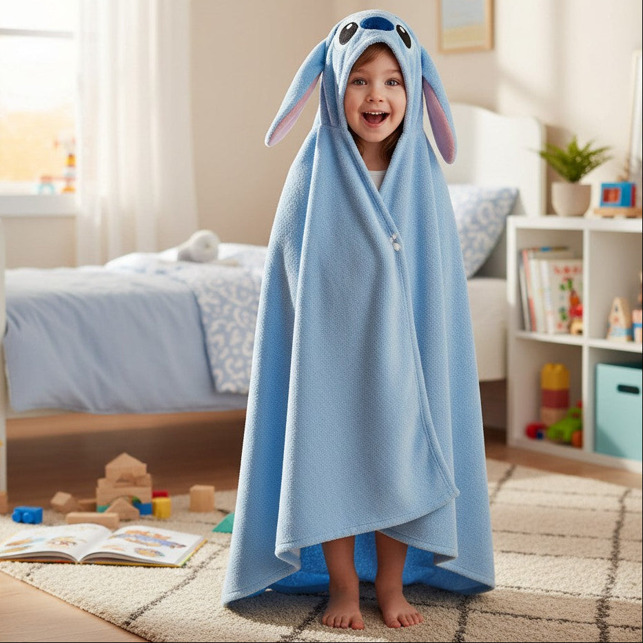 A young child stands barefoot in a bright bedroom, wrapped in a light blue hooded blanket with long bunny ears and a stitched face design.