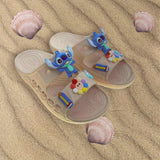 Beige double strap slides fitted with Stitch and rainbow charms resting on soft sand