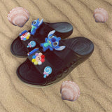 Black foam slides with Stitch, flower and candy charms laid on rippled sand