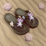 Dark brown thong sandals topped with purple and pink character charms on rippled sand