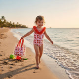 Girl walking on the beach in a strawberry and heart print onepiece swimsuit with a tote bag