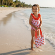 Girl in a strawberry and heart print onepiece swimsuit standing at the shoreline with a tote bag