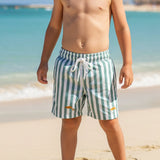 Young boy in striped swim shorts standing with small fish on a sandy beach
