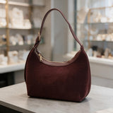 Burgundy suede finish hobo bag with a buckle strap and gold zip top on a stone counter
