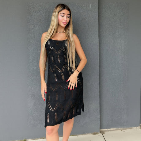 A woman modeling a sleeveless black crochet midi dress with geometric openwork patterns standing against a textured grey wall