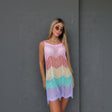 A woman modeling a sleeveless crochet knit mini dress with a pastel chevron pattern in purple, pink, peach, cream, and turquoise, standing against a dark grey textured wall