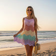 A woman stands on a sandy beach at sunset wearing a sleeveless crochet mini dress with a pastel chevron pattern in pink, orange, yellow, green, and purple