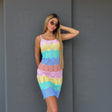 A woman modeling a pastel striped crochet mini dress with horizontal bands of pink, yellow, teal, blue, and peach, posing against a dark grey textured wall