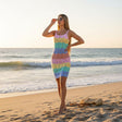 A woman stands on a sandy beach at sunset wearing a sleeveless, bodycon knit dress with horizontal pastel stripes in pink, yellow, green, blue, and red