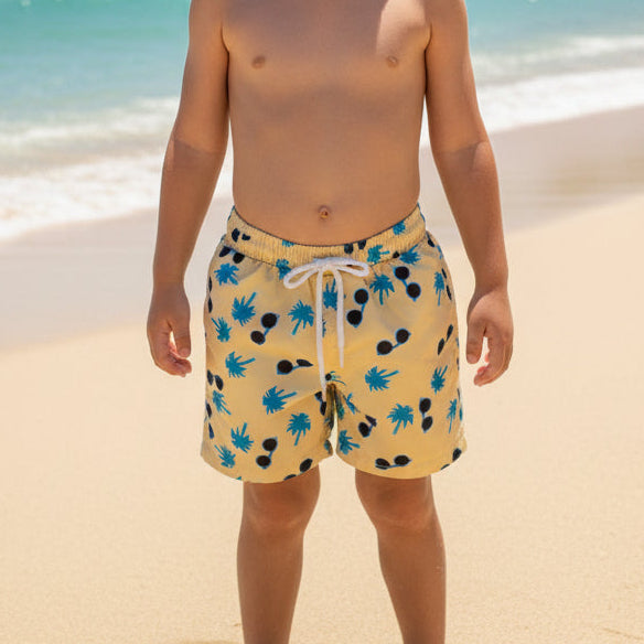 Child wearing yellow swim shorts with blue palm trees and sunglasses on a beach