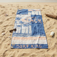 A rectangular beach towel laid flat on a sandy beach near the shoreline, featuring a blue and white tropical palm tree design with the words “A GALLEFORD SURF KING” and “SURF KING.”.