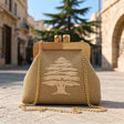 Tan woven clutch with a cream cedar tree, wooden frame clasp and gold chain on an old stone street