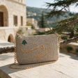 Speckled taupe pouch on a gold chain, set on a stone wall beneath pine needles with an old sandstone house behind