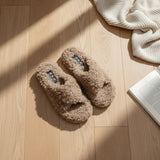 Taupe boucle crossover slippers laid flat on a sunlit wooden floor beside a knit throw