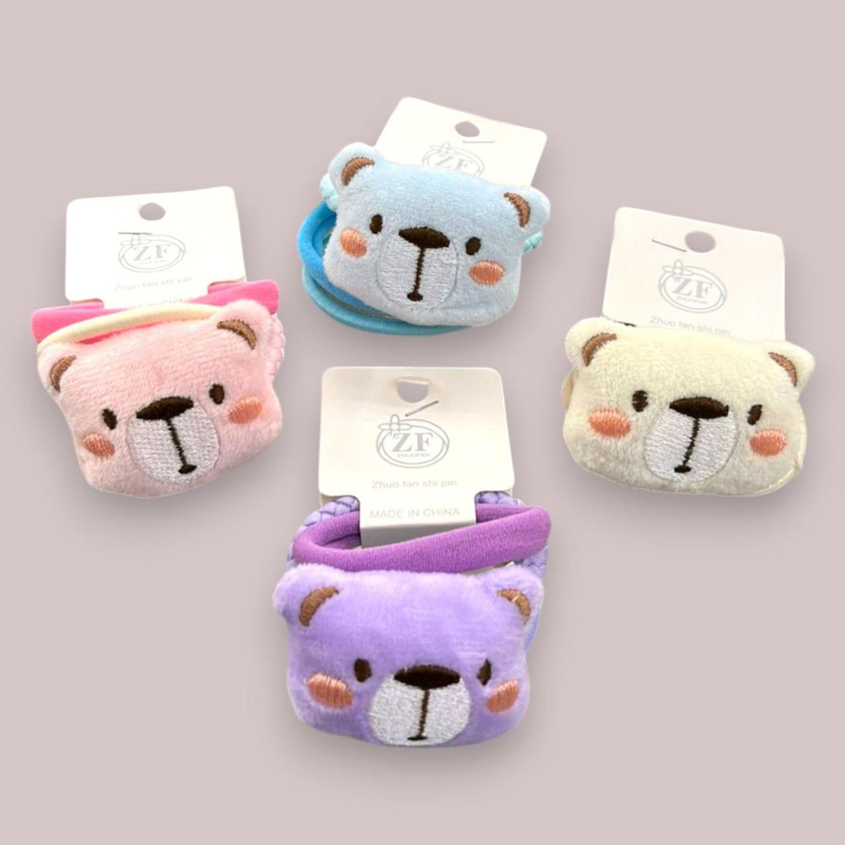 Four plush teddy bear face hair ties in pink, blue, cream and purple with embroidered muzzles and rosy cheeks