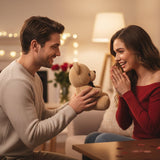 Man kneeling to offer the caramel bow bear to a delighted woman on a sofa
