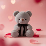 Grey plush bear with a black velvet bow on a pink backdrop scattered with rose petals