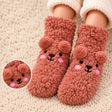 Child wearing terracotta fuzzy socks with a smiling bear face, pom pom ears and pink cheeks