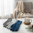 Three pairs of gloves in dark-grey, light-grey, and blue on a table with a mug of hot chocolate and festive decorations