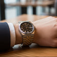 Close-up of a wrist wearing a silver watch with a brown dial on a wooden surface