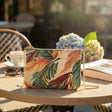 Tropical leaf tapestry crossbody bag with a gold chain on a cafe table