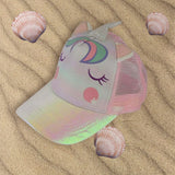 Iridescent unicorn cap with a mesh back laid on sand with shells
