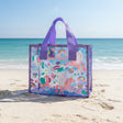 Purple glitter trimmed clear tote covered in winged unicorns and rainbows, sitting on beach sand