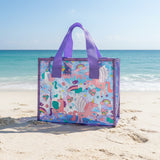 Purple glitter trimmed clear tote covered in winged unicorns and rainbows, sitting on beach sand