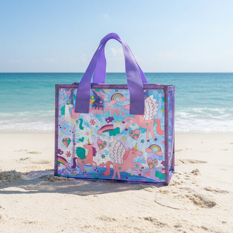 Purple glitter trimmed clear tote covered in winged unicorns and rainbows, sitting on beach sand