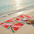 Beach towel with watermelon pattern on a sandy beach with ocean waves.