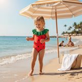 Child in a watermelon-themed swimsuit standing on a sandy beach with an umbrella and beach bag in the background.