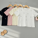 Four t-shirts in black, pink, beige, and white with heart patterns on hangers against a gray background.