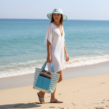 Woman in a white dress and blue hat holding a striped bag on a beach.