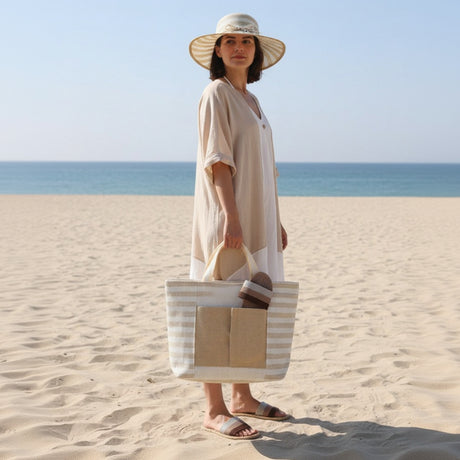 Woman on a beach wearing a beige hat and dress, holding a striped tote bag.