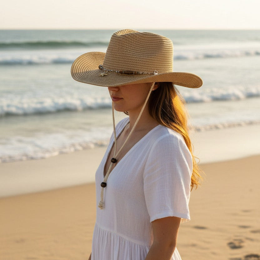 Woman in a white dress and straw hat standing on a beach with ocean waves in the background