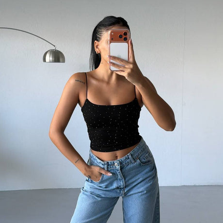 Person wearing a black top and blue jeans taking a mirror selfie against a plain wall.