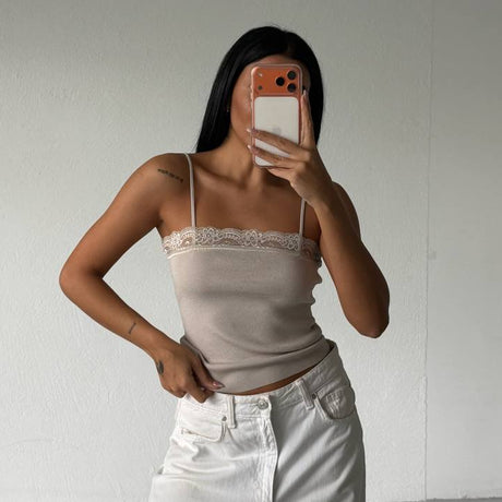 Person wearing a beige top and white pants taking a mirror selfie against a plain white wall.